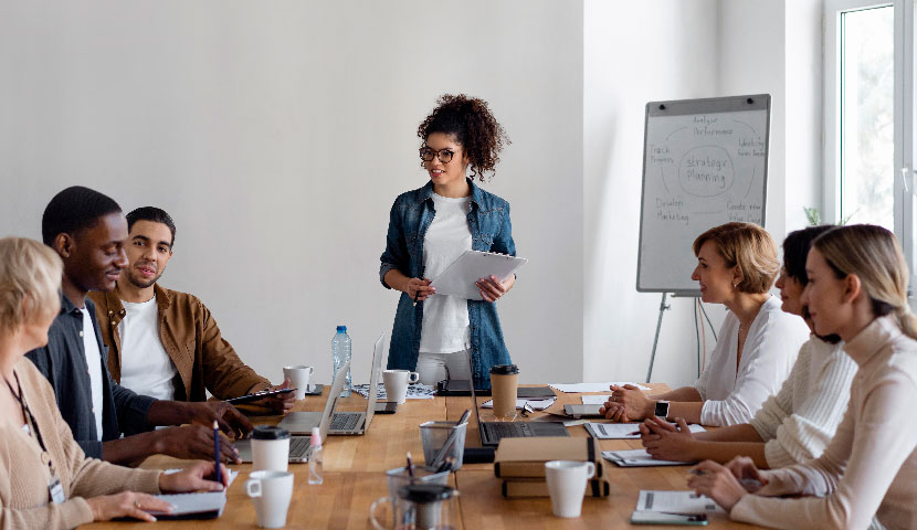 Grupo de personas en una sala de reuniones, sentados alrededor de una mesa con laptops y documentos. Una mujer de pie sostiene una tableta y parece estar presentando o liderando la reunin, mientras los dems la escuchan. En el fondo, hay un rotafolios con